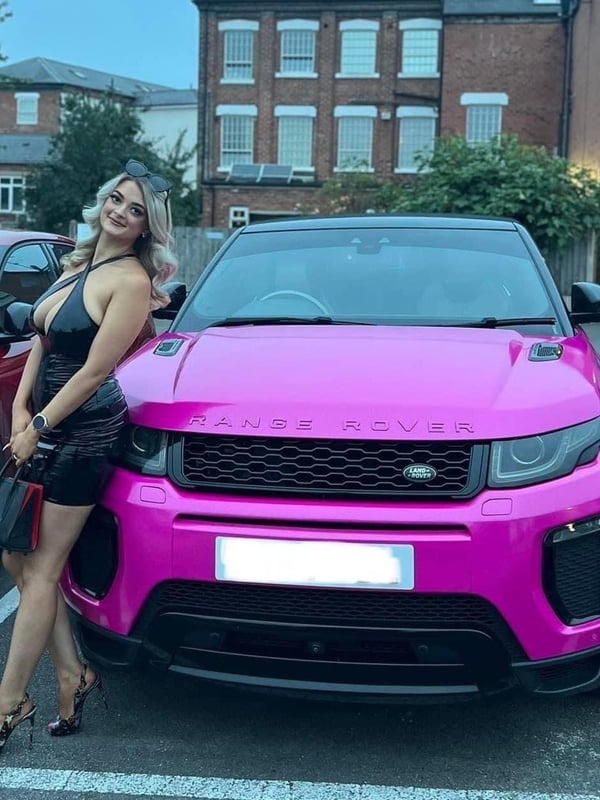 Young woman with blonde hair wearing a black dress and high heels, posing next to a pink Range Rover car parked outside brick buildings in an evening setting.