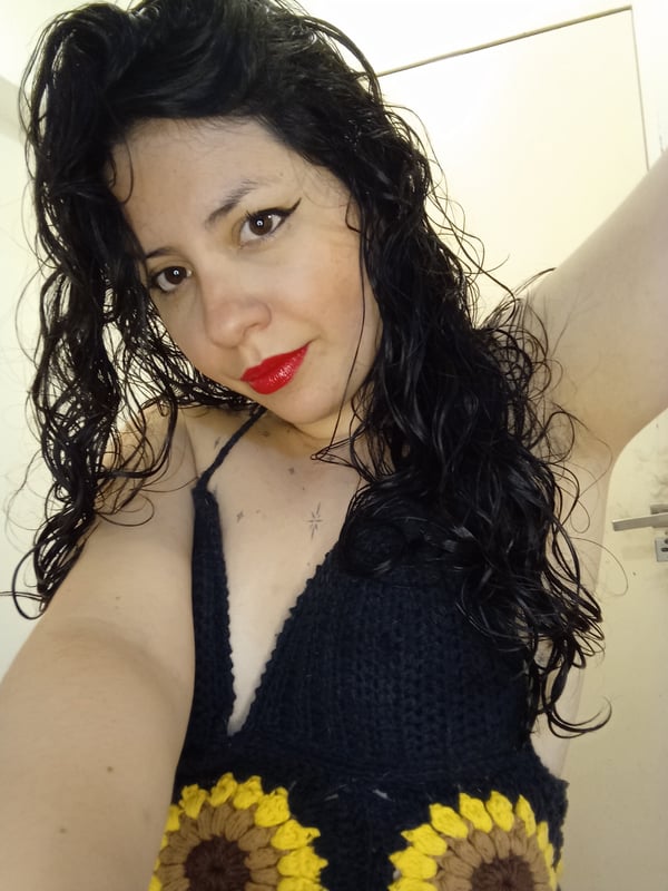 A woman with long dark curly hair smiling at the camera, wearing a black knitted top with yellow sunflower designs.
