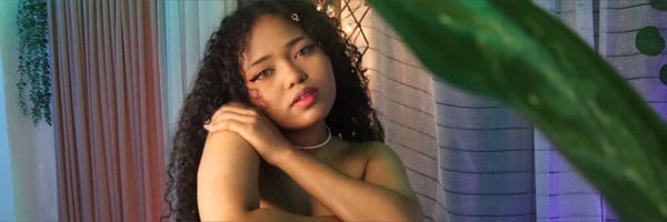 A young woman with curly hair posing confidently in a colorful indoor setting with plants and draped fabrics.