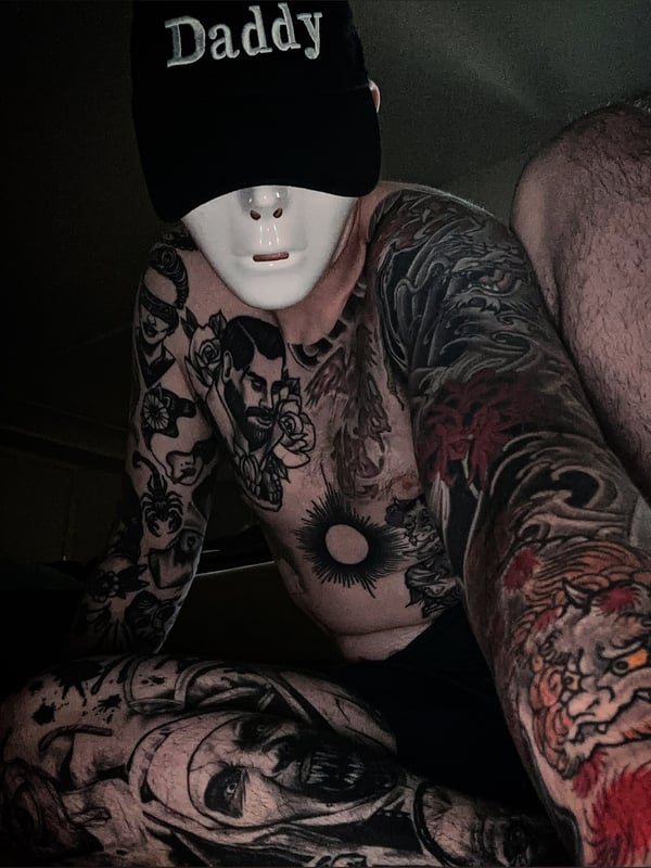 Person wearing a black 'Daddy' cap and white mask, displaying extensive colorful tattoos on arms and chest while seated.