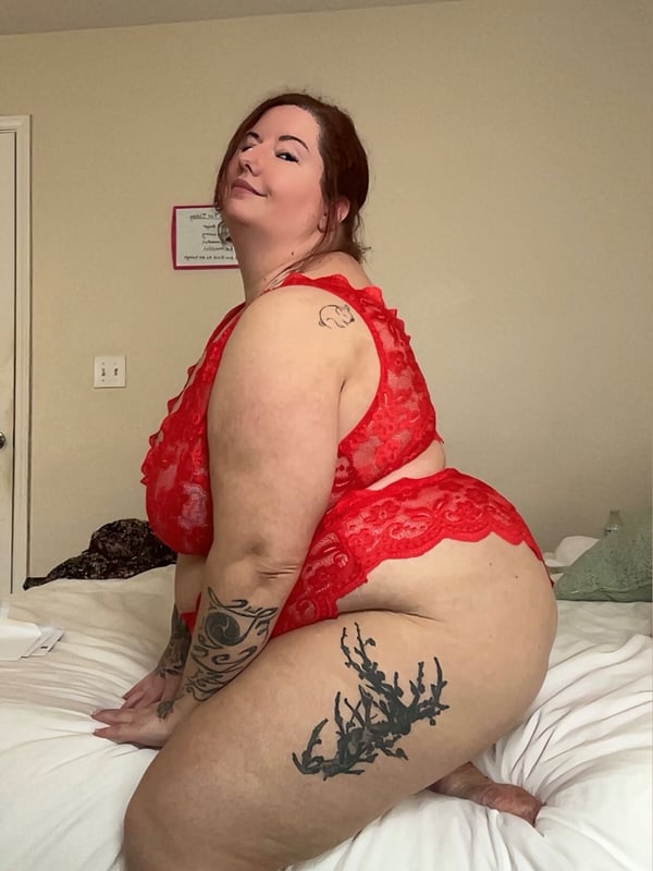 A woman in red lace lingerie sitting on a bed, smiling with tattoos visible on her arms.