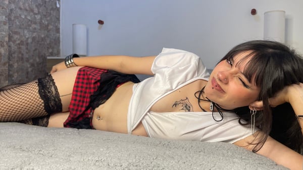 Young woman lounging on a bed in a plaid skirt, fishnet stockings, and a cropped white top, posing playfully in a cozy bedroom setting.