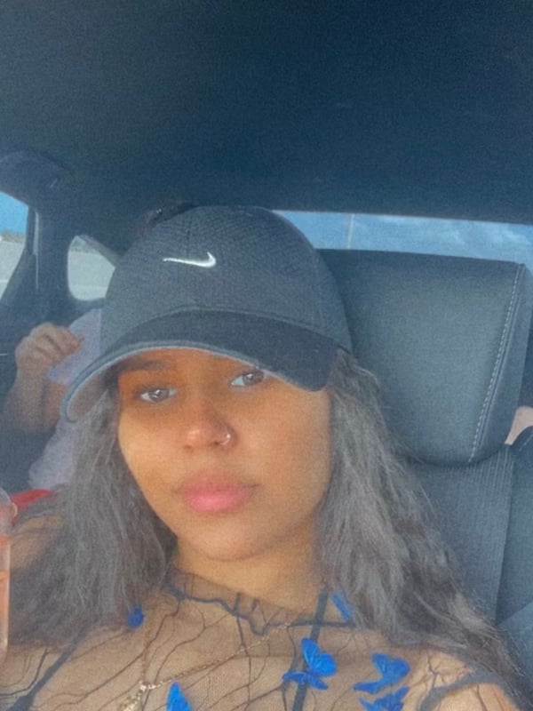 A young woman wearing a cap and mesh top, sitting in a car and smiling at the camera.