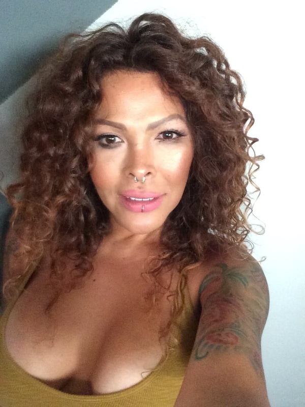 Close-up selfie of a woman with curly hair, smiling at the camera, wearing a sleeveless top, with tattoos visible on her arm.