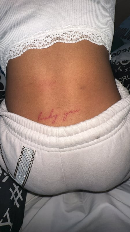 A person standing with back turned, wearing a white lace-trimmed top and gray sweatpants, showing a small tattoo on the lower back.