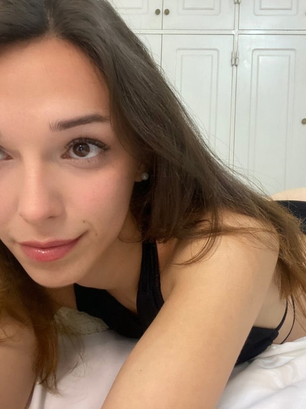 Close-up of a young woman with long dark hair and subtle makeup, lying on a bed facing the camera, with wooden cabinets in the background.