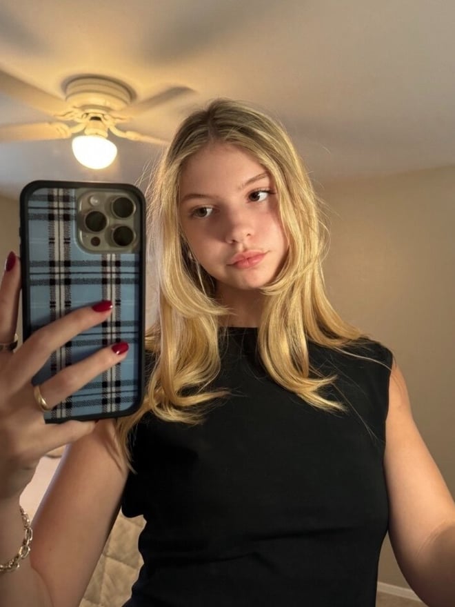 A blonde woman taking a mirror selfie indoors while wearing a black top.