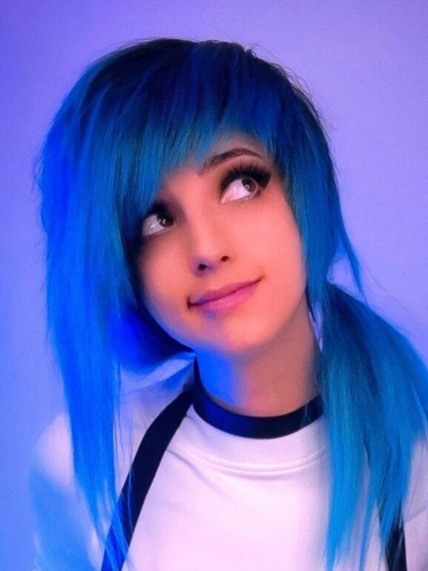 A young woman with bright blue hair in pigtails, wearing a white top with black choker, posing playfully against a purple background.