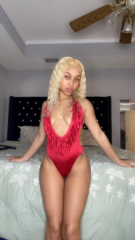 A woman posing confidently on a bed in a red fringed outfit inside a modern bedroom with ceiling fan and neutral decor.