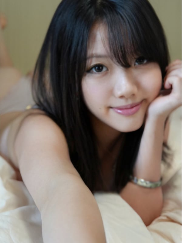 A young woman with straight black hair and bangs smiling at the camera, resting her chin on her hand while lying down.