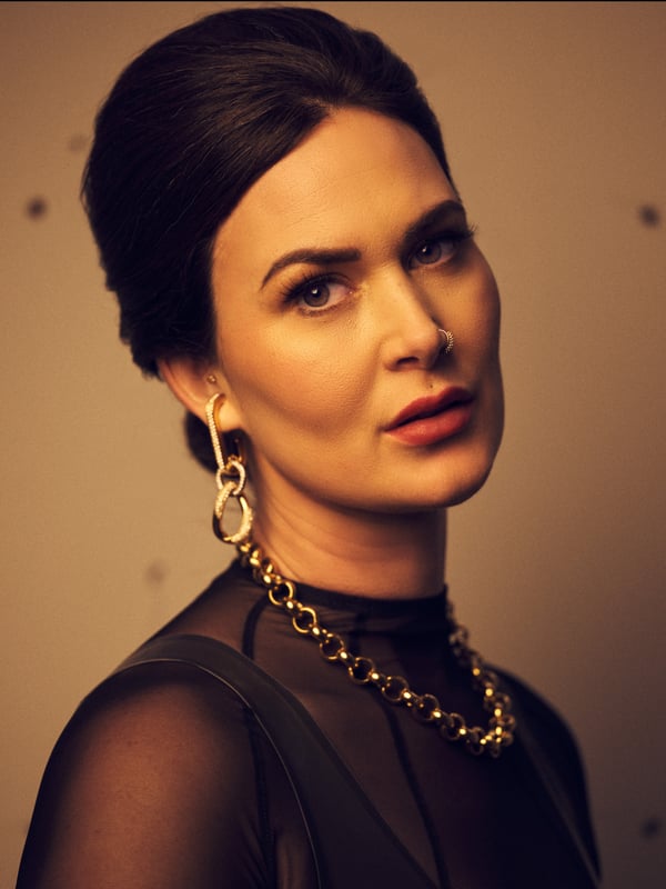 A woman with brunette hair in an updo, wearing a sheer black top and gold jewelry, gazing directly at the camera against a neutral background.