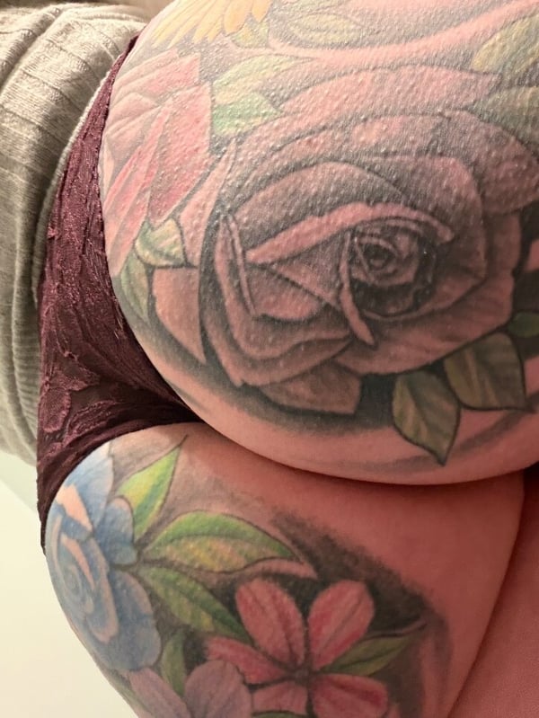 Close-up view of a person's lower body with colorful floral tattoos on the skin, wearing soft gray and maroon clothing against a neutral background.