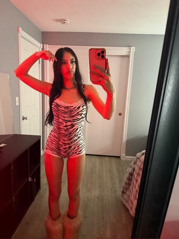 Young woman with long dark hair posing in a zebra-print bodysuit and fluffy boots, taking a mirror selfie in a bedroom with warm lighting.
