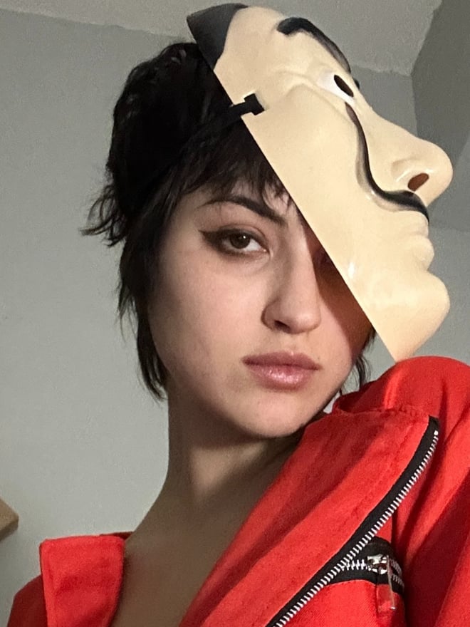 Close-up of a young woman with short dark hair and dramatic makeup wearing a red jumpsuit, holding a white mask over part of her face.