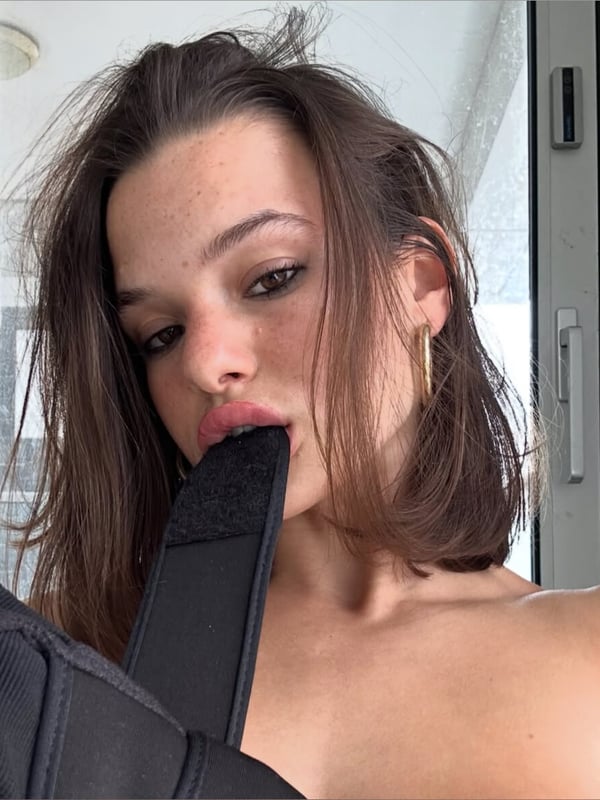 Close-up of a young woman with shoulder-length brown hair, wearing hoop earrings and posing with a black strap near her face indoors.