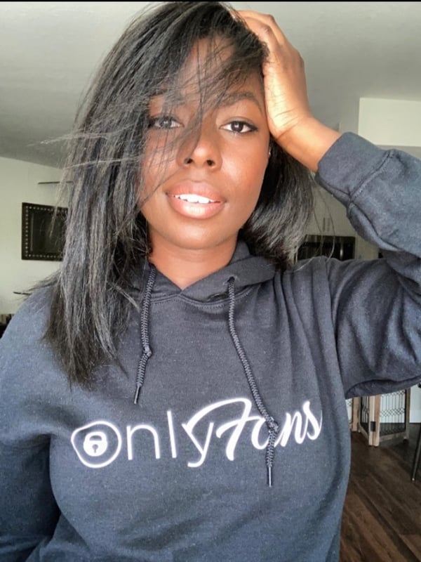A woman with long dark hair smiling while wearing a gray OnlyFans hoodie indoors.