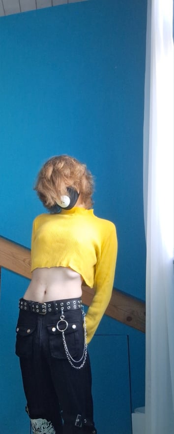 A person with curly orange hair wearing a yellow crop top and black pants with chains, posing in front of a blue wall.