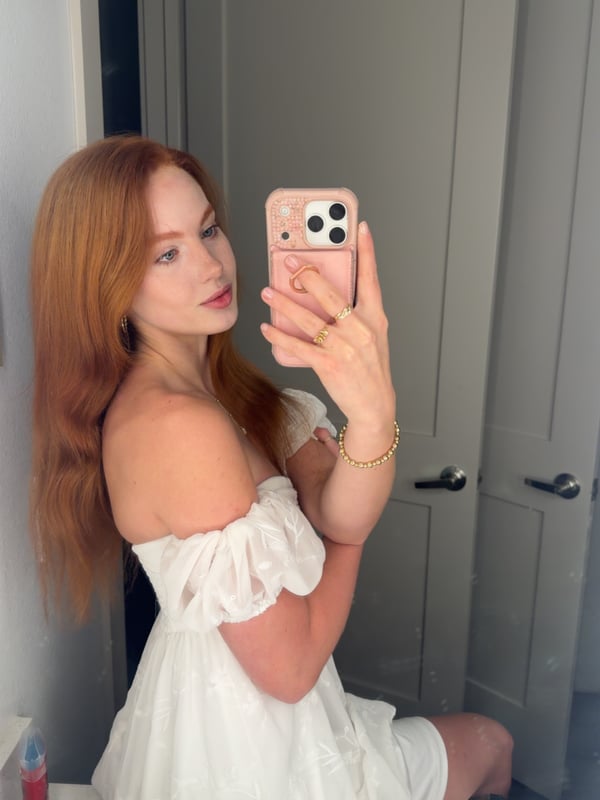 Young woman with blonde hair taking a selfie in a black halter top inside a bedroom with warm lighting.