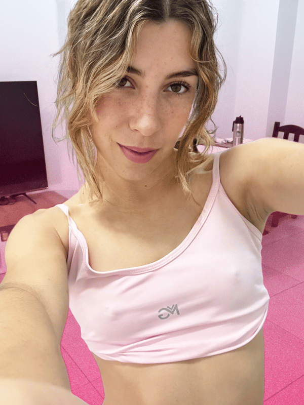 A young woman with light brown hair taking a selfie indoors while wearing a pink tank top.