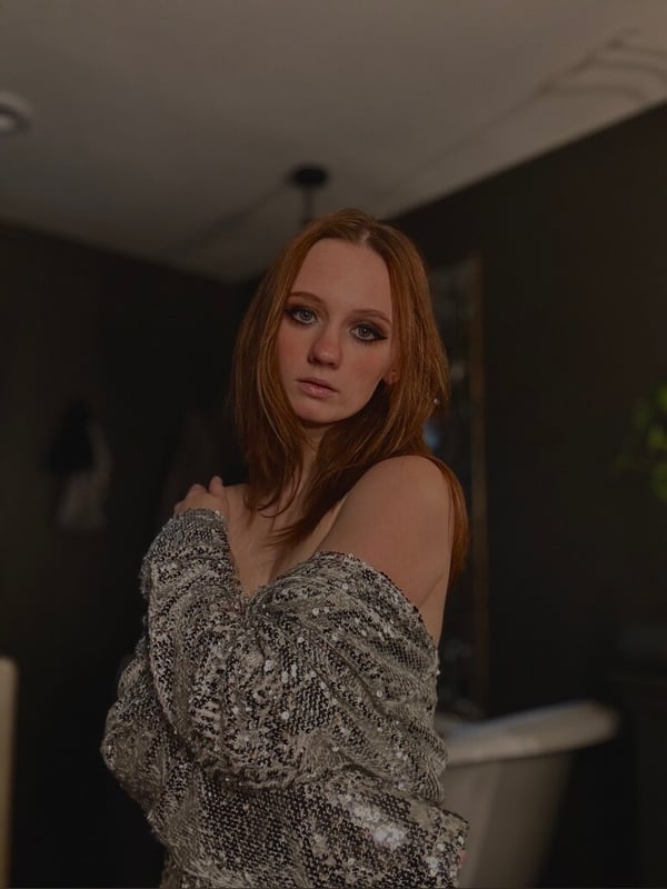 A woman with long red hair poses thoughtfully in a sparkly off-shoulder top inside a cozy room with a mirror and houseplant.