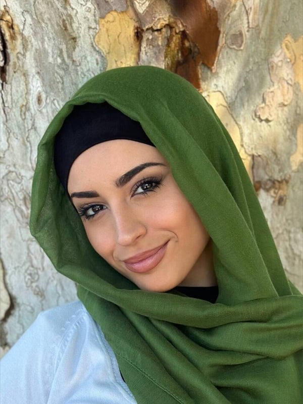 Close-up portrait of a smiling woman in a green hijab against a tree trunk background.