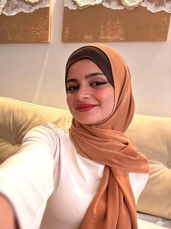 A woman wearing a hijab and white top smiles at the camera in a selfie, seated against a neutral background with decorative wall art.