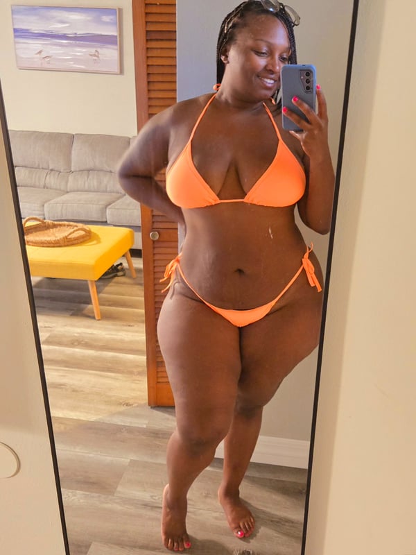 A woman taking a mirror selfie in an orange bikini, standing in a cozy room with wooden floors and beach artwork on the wall.