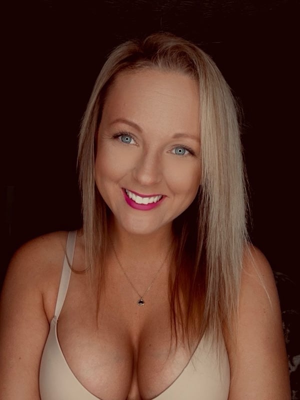 A smiling young woman with long blonde hair, wearing a beige bra and necklace, posing against a dark background.