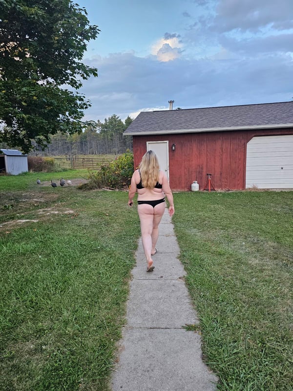 A woman with long blonde hair walks along a path in a rural farm setting with a red barn, trees, and open fields under a cloudy sky.
