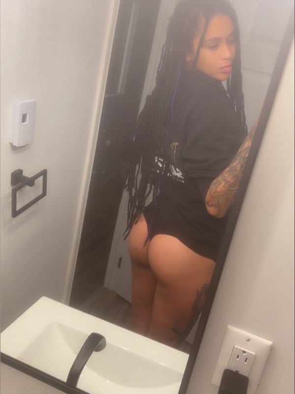 A woman with long braided hair standing in a bathroom mirror, wearing a black top and shorts.