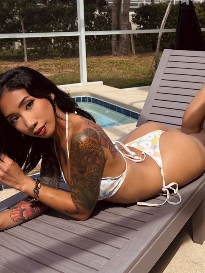 A tattooed woman in a floral bikini reclining on a poolside lounge chair.
