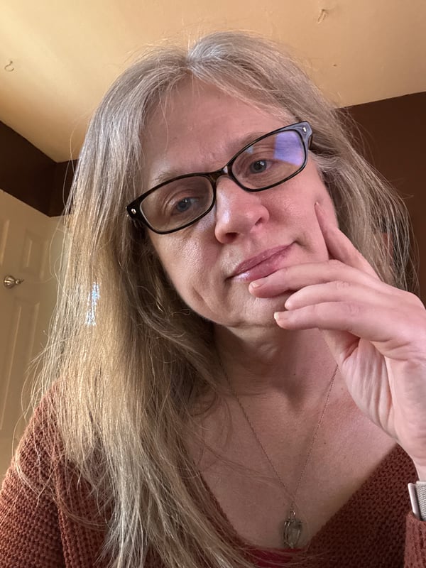 A woman with blonde hair and glasses wearing a knitted top, posing thoughtfully with her hand on her chin against a warm indoor background.