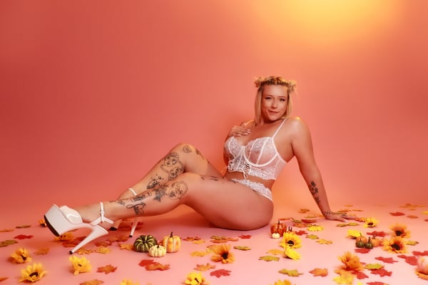 Woman with blonde hair wearing a white lingerie set and high heels, posing on a pink background surrounded by autumn leaves and pumpkins.
