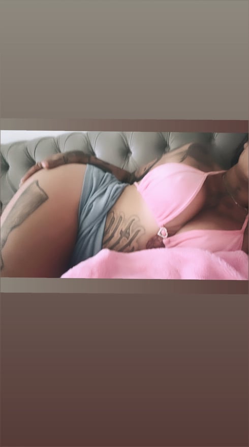 A woman reclining on a couch in a pink top and shorts, with tattoos visible on her leg and midriff.