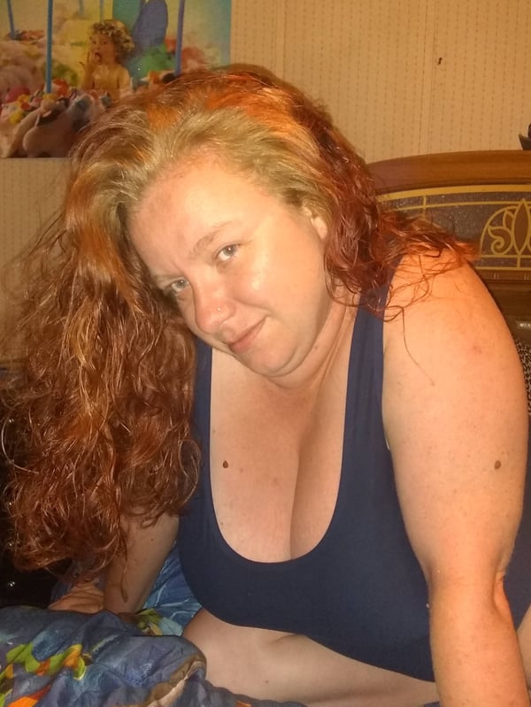 Woman with strawberry blonde hair smiling while leaning forward in a tank top inside a cozy bedroom with colorful artwork and wooden furniture.
