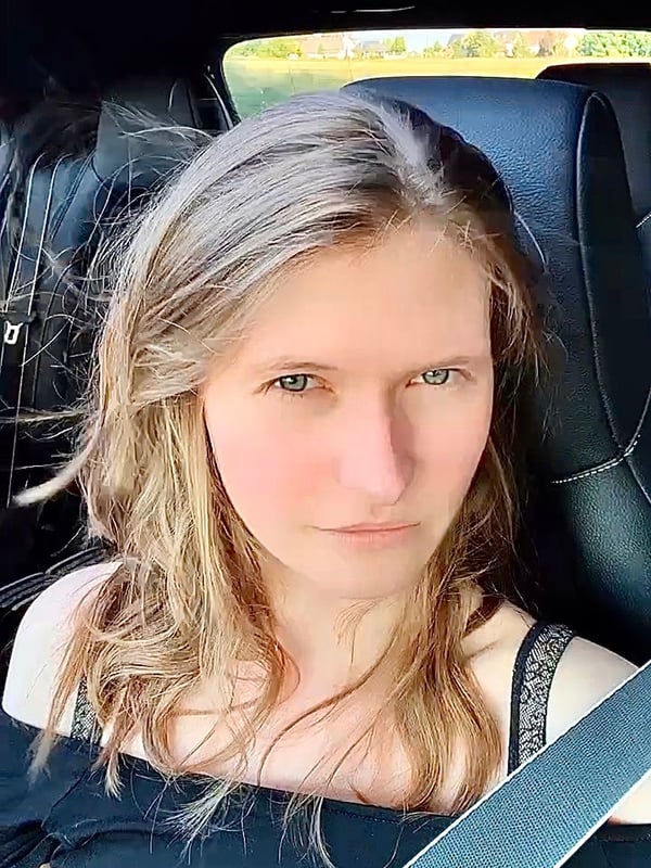 Young woman with long wavy blonde hair sitting in a car, wearing an off-shoulder top and seatbelt, gazing directly at the camera.