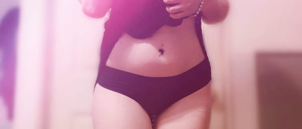 A person posing in dark lingerie with a visible navel piercing in a softly lit indoor setting.