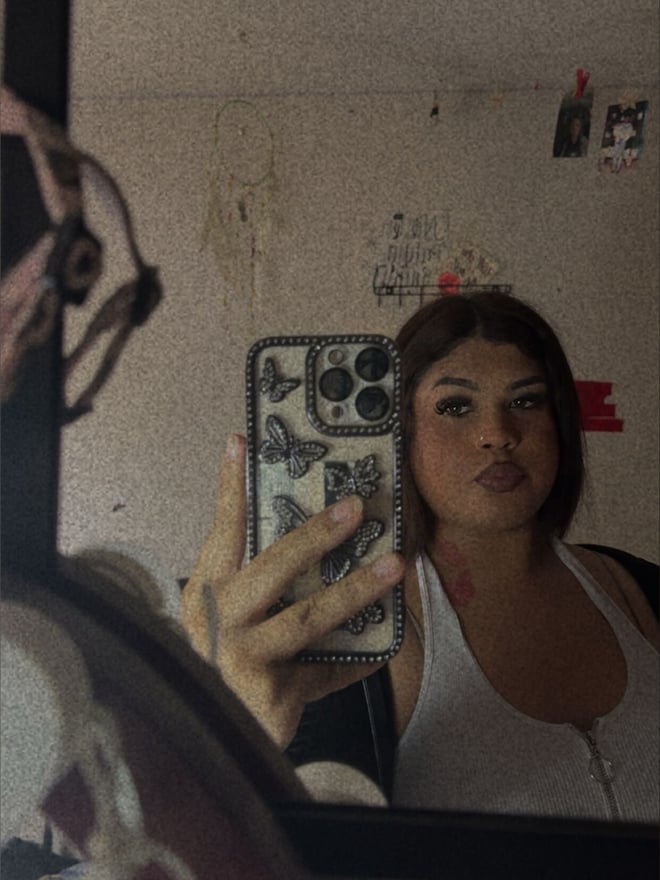 Young woman taking a mirror selfie in a bedroom, wearing a white tank top and holding her phone, with subtle makeup and brown hair.