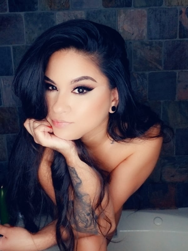 A woman with long dark hair posing confidently in a bathroom setting with tattoos on her arm.