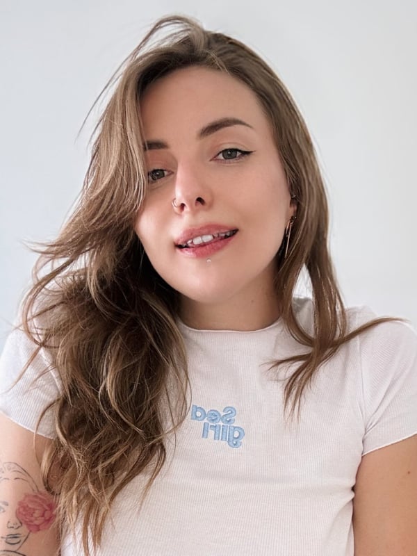 A young woman with wavy brown hair smiling playfully at the camera, wearing a white t-shirt with text, visible tattoos on her arm.