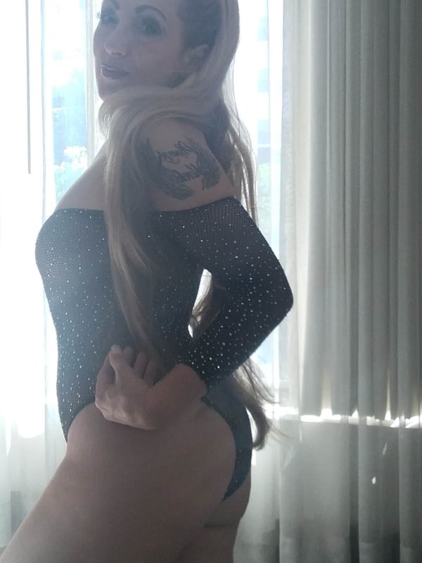 Woman posing confidently in a sparkling black bodysuit near a window with sheer curtains, highlighting her tattoos and long blonde hair.