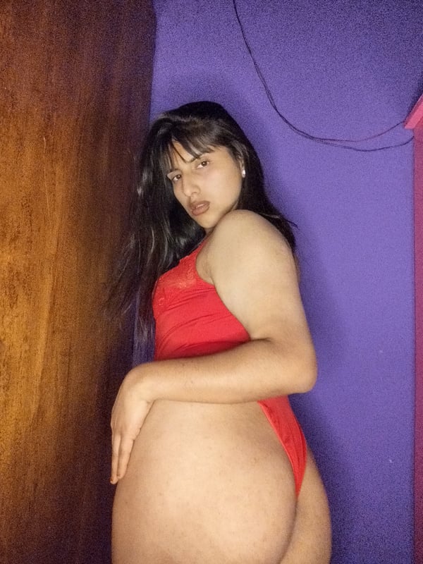 Young woman with long dark hair standing sideways in a red swimsuit against a purple wall.