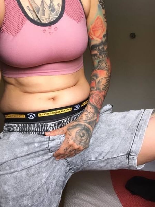 A tattooed woman in a pink sports bra and gray shorts stands confidently in a casual indoor setting.