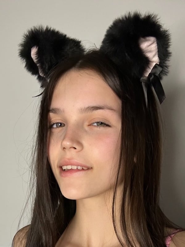 Close-up portrait of a young woman with long dark hair and subtle makeup, wearing black cat ear headbands against a plain background.