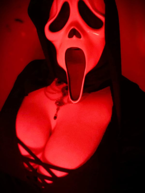Person wearing a red-tinted Scream mask and dark hooded outfit, posing with a necklace visible.