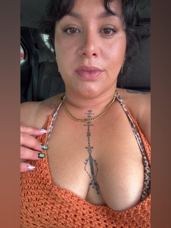 Woman with curly dark hair and tattoos on her chest, wearing a low-cut orange top and gold necklace, posing in a car.
