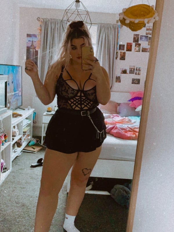 A woman taking a mirror selfie in a bedroom, wearing a black lace top and shorts with white socks, surrounded by casual room decor and hanging lights.