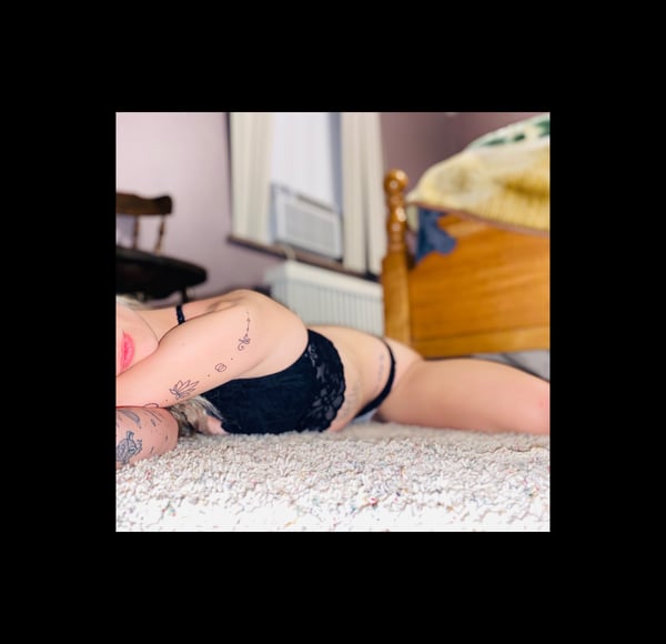 A woman lounging on the floor in a bedroom, wearing lace lingerie and posing near a bed with wooden accents.