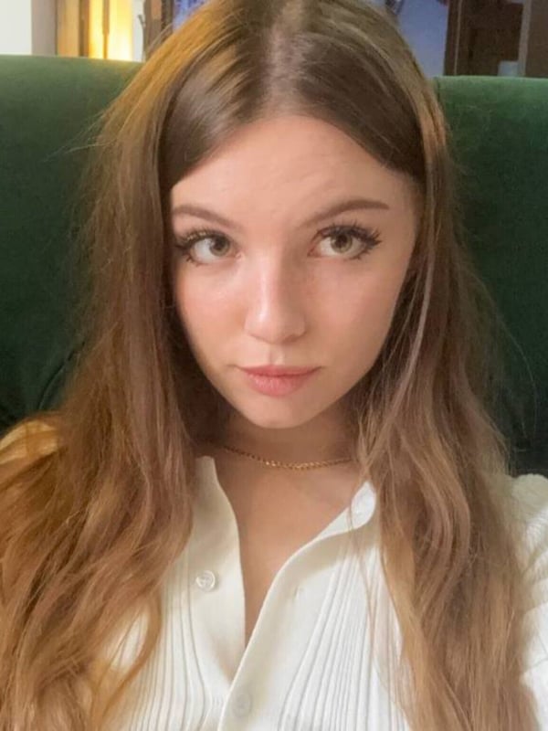 Young woman with long brown hair sitting on a green chair, wearing a white shirt, with a neutral expression.