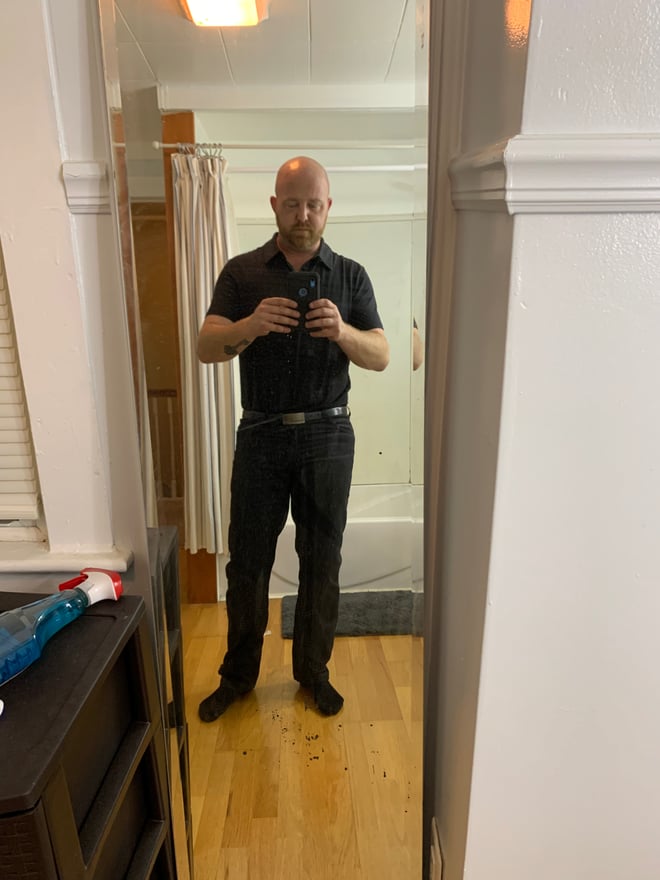 A bald man in a black shirt and pants standing in a bathroom, taking a mirror selfie.